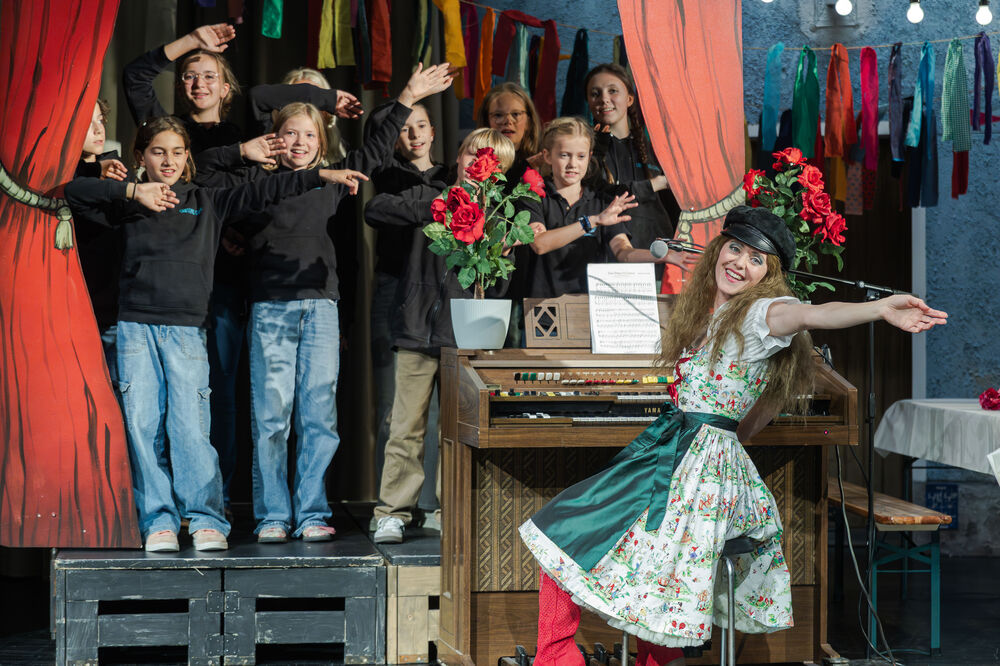 Im Vordergrund dreht sich eine Person in einem geblümten Dirndl mit ausgebreiteten Armen von einer Orgel weg Richtung Kamera. Im Hintergrund steht ein Kinderchor mit erhobenen Armen auf einer Bühne.