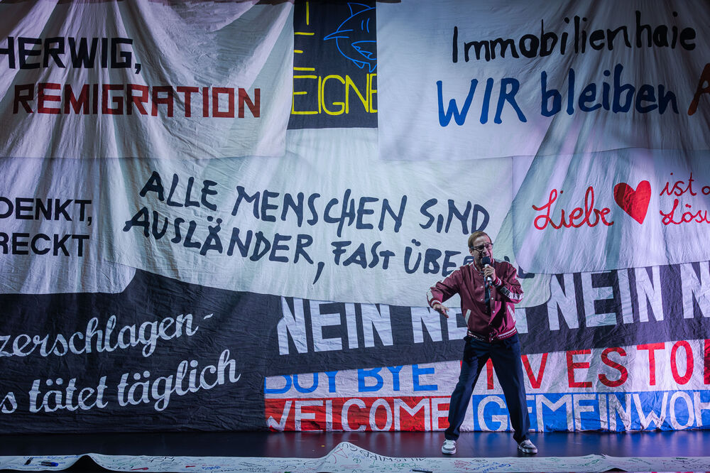 Eine kurzhaarige Person mit Brille und sportlicher Jacke spricht angeregt in ein Mikrofon, ähnlich wie ein Sportjournalist. Im Hintergrund hängen überlappende Banner; auf einem ist deutlich der Schriftzug zu lesen: ‚Alle Menschen sind Ausländer, fast überall‘.